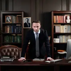 Poważny adwokat w garniturze stoi przy biurku w gabinecie. Zastanawia się, ile zarabia adwokat w Warszawie, otoczony książkami i flagą USA.