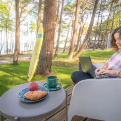 Kobieta pracuje na laptopie przy stoliku z kawą i croissantem. W tle widać morze i sosny, idealne miejsce dla domków z wyjściem na plażę.