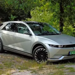 Srebrny Hyundai Ioniq 5 z czarnymi felgami stoi na kamienistej drodze, otoczony zielenią i starym budynkiem.