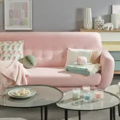 Sofá rosa pastel con cojines y mantas, mesas de centro con velas y malvaviscos, y un mueble de cajones de colores.