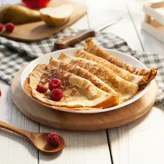 Frisch zubereitete Pfannkuchen, ein köstliches Pancakes Rezept, garniert mit Himbeeren auf einem Teller. Daneben liegen Birnen und ein Holzlöffel.