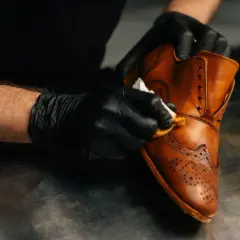 Mains gantées lustrant une chaussure en cuir marron, un soin méticuleux pour un cuir machine à laver qui retrouve son éclat.