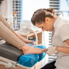 Profesjonalny pedicure co to? Kosmetyczka w białej bluzce i rękawiczkach dba o stopy klientki na wygodnym fotelu.