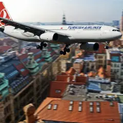 Samolot Turkish Airlines przelatuje nad miastem, jakby zmierzał w kierunku **Zakaukazia**. W tle widać zabytkowe budynki z czerwonymi dachami.