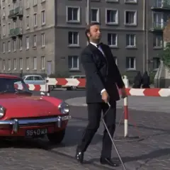 Mężczyzna w garniturze z laską idzie obok czerwonego samochodu. Chyba nie lubię poniedziałku, bo mam ochotę uciec tym autem.