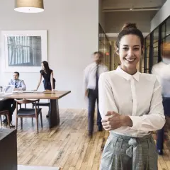 Eine lächelnde Frau steht im Vordergrund, während im Hintergrund verschwommene Kollegen arbeiten. Sie repräsentiert die Dynamik von IU Kommunikation und PR.