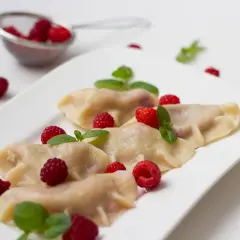 Pyszne pierogi z malinami, udekorowane świeżymi malinami i listkami mięty, prezentują się apetycznie na białym talerzu.