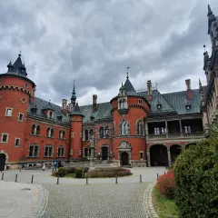 Pławniowice atrakcje: okazały zamek z czerwonej cegły z wieżami i arkadowym gankiem, otoczony brukowanym dziedzińcem i zielenią.