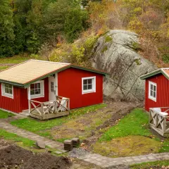Casitas rojas con tejados naranjas y amarillos, rodeadas de un suelo rústico con hierba y tierra removida, bajo un gran peñasco y árboles otoñales.