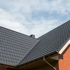 Szary dach z blachy modułowej, z widocznymi rynnami i fragmentem ceglanej ściany. Idealne wymiary dla każdego domu.