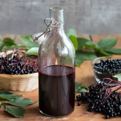Butelka z ciemnym sokiem z czarnego bzu, obok koszyk z owocami i miseczka z jagodami.