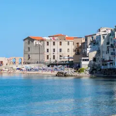 Edificios antiguos y una playa animada en Cefalú, Italia, con el mar azul brillante.