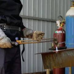 Uczestnik kursu spawania gazowego precyzyjnie tnie metalowy element palnikiem. W tle widać butle z gazami.