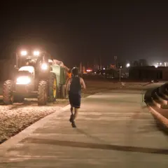 Mężczyzna uprawia bieganie w nocy na promenadzie, obok pracującego traktora na plaży.