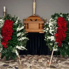 Dwa wieńce pogrzebowe z czerwonych róż i białych kwiatów stoją obok drewnianej trumny. Czy na kremację kupuje się kwiaty? Tak, to piękny gest.