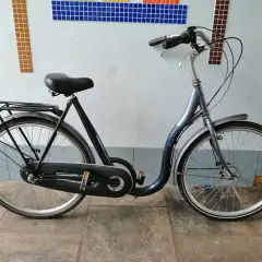 Donkerblauwe cruiser fiets met bagagedrager, geparkeerd voor een muur met mozaïek.