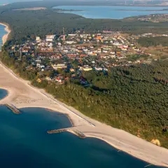 Jarosławiec atrakcje: aerial view of a coastal town with sandy beaches, breakwaters, pine forests, and a lake in the distance.