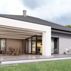 Nowoczesna dobudówka do domu z tarasem i meblami ogrodowymi. Jasna elewacja, duże okna i oświetlenie tworzą przytulną atmosferę.
