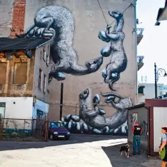 Niesamowite street art przykłady: wielkie, futrzaste zwierzęta bawią się jajkami na ścianie kamienicy. Obok przechodzą ludzie z psem.