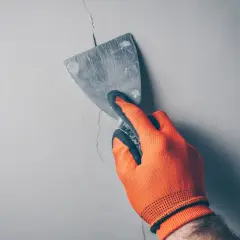 Ręka w pomarańczowej rękawicy wypełnia głębokie pęknięcia w ścianie szpachlą za pomocą kielni.