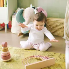 Ein **baby 9 monate** sitzt auf einem Teppich und spielt mit Holzspielzeug. Im Hintergrund sind Stofftiere und ein Kinderbett zu sehen.