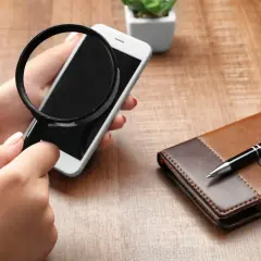 Dłoń trzyma lupę nad ekranem telefonu, jakby szukała ukrytych informacji. Obok leży skórzany notatnik z długopisem.