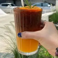 Orzeźwiające espresso z sokiem pomarańczowym, ozdobione plasterkiem pomarańczy i gałązką rozmarynu, w dłoni z kolorową bransoletką.