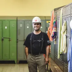 Ein Mann in Arbeitskleidung und Helm steht in einer Umkleidekabine. Handtücher hängen an den Spinden. Die Umziehzeit ist Teil der Arbeitszeit.