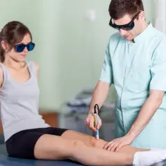 Fizjoterapeuta wykonuje laser rehabilitację na kolanie pacjentki. Oboje noszą okulary ochronne.