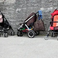 Trzy puste wózki dziecięce stoją przy kamiennej ścianie. Jeden ma torbę w kwiaty, drugi brązową torbę i niebieską chustę, a trzeci pomarańczowy kocyk.