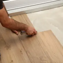 A person installs wood-look drop and lock flooring, using a tool to secure a plank onto adhesive-covered subfloor.