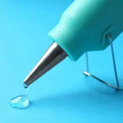Un pistolet à colle turquoise dépose une goutte de colle transparente sur un fond bleu. Une petite tache de colle se forme.
