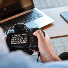 Ręce trzymają aparat, na ekranie ustawienia manualne. Nauka fotografii w praktyce, obok laptop i telefon.