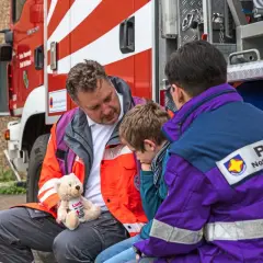 Ein Mann mit einem Teddybär spricht mit einem Kind neben einem Feuerwehrauto. Eine Person mit PSNV-Aufschrift auf der Jacke sitzt daneben.