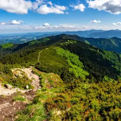 Widok z **przełęczy Karkoszczonka** na zielone, zalesione góry i błękitne niebo z chmurami. Szlak turystyczny wije się po zboczu.