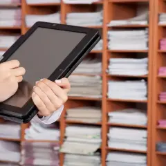 Une personne utilise une tablette pour gérer des documents, illustrant l'archivage électronique, une obligation légale. Les étagères remplies de dossiers rappellent l'importance de la numérisation.