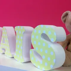 Lettre en relief "THAIS" décorée de pois sur fond rose vif, à côté d'un ours en peluche.