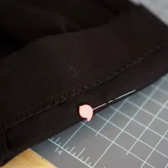 Close-up of black pants hem, pinned and ready for an invisible stitch. A pink pin secures the fabric on a cutting mat.