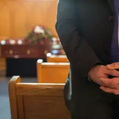 Mężczyzna w garniturze stoi w kaplicy pogrzebowej, czekając na rozpoczęcie ceremonii. W tle widoczna trumna i ławki. Zasiłek pogrzebowy KRUS może pomóc w pokryciu kosztów.