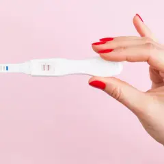 Dłoń z czerwonymi paznokciami trzyma test ciążowy z dwoma kreskami, sugerującymi ciążę biochemiczną.