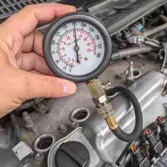 Mężczyzna sprawdza kompresję silnika za pomocą manometru. Wskaźnik pokazuje ok. 170 psi.