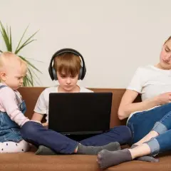 Dzieci na kanapie, chłopiec w słuchawkach gra na laptopie, maluch obok. Matka z telefonem. Kontrola rodzicielska w Polsce trwa do 18 lat.