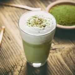 Szklanka z zielonym matcha latte z pianką, obok miseczka z proszkiem matcha i bambusowa trzepaczka.
