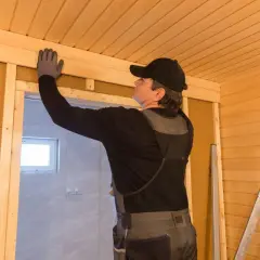 Budowa sauny na poddaszu. Mężczyzna w rękawiczkach montuje drewniane wykończenie sufitu.