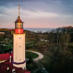 Najstarsza latarnia morska w Polsce, wysoka, kremowa wieża z czerwonymi akcentami, góruje nad krajobrazem o zachodzie słońca.