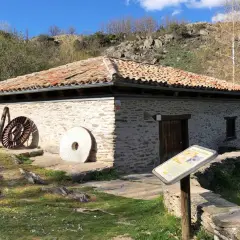 El molino de la Hiruela, con su tejado de tejas rojas y muros de piedra, se alza junto a un río. A su lado, piedras de moler y una rueda de hierro evocan su pasado.