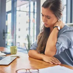 Kobieta z bólem karku przy biurku, pierwszy raz odczuwa taki dyskomfort.