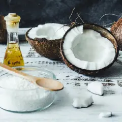 Coupé en deux, un coco frais et sa chair blanche côtoient une petite fiole d'huile dorée. Un bol de noix de coco râpée et une cuillère en bois suggèrent l'utilisation de l'huile de coco pour des recettes gourmandes ou des soins.