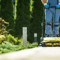 Pracownik w niebieskim kombinezonie używa **zamiatarki do liści**, aby oczyścić ścieżkę w pięknym ogrodzie.