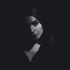 A woman's face emerges from darkness, her features dramatically sculpted by side lighting. A subtle nose ring catches the light.